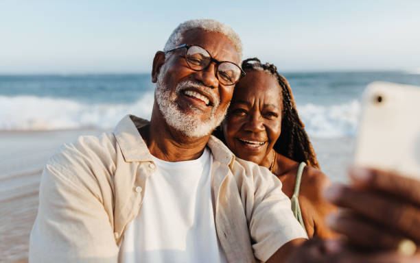 An older African American couple shares a joyful moment, taking a selfie together with the ocean backdrop during a serene beach walk.
