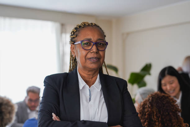 Portrait of senior african woman looking on camera inside business office with colleagues in background - Entrepreneurs people and finance concept - Focus on face